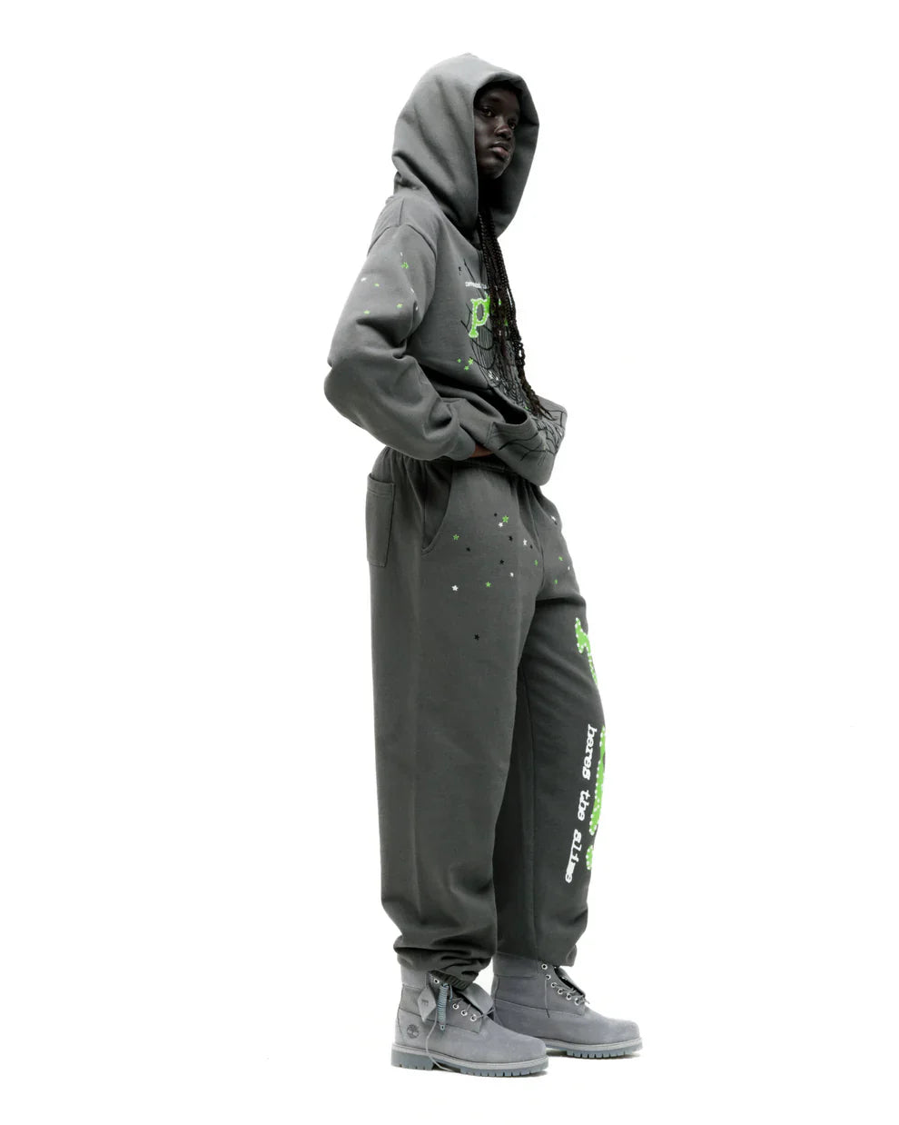 A person wears the Sp5der “P!nk! Hares the Slime” Hoodie – Olive Green 440GSM Heavyweight Spider-Web Pullover, standing sideways with hands in pockets and hood up, paired with sweatpants and gray boots against a white background.