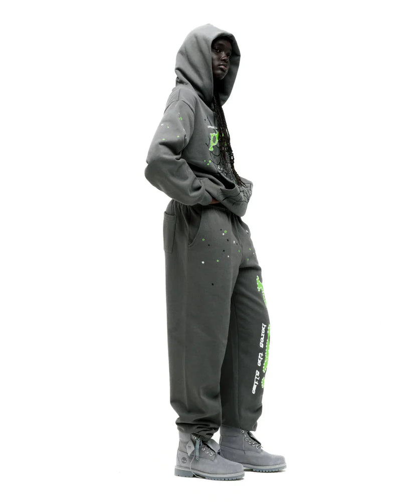 A person wears the Sp5der “P!nk! Hares the Slime” Hoodie – Olive Green 440GSM Heavyweight Spider-Web Pullover, standing sideways with hands in pockets and hood up, paired with sweatpants and gray boots against a white background.