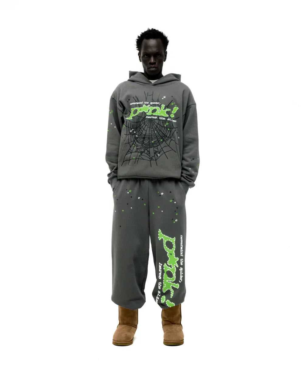 A person stands against a white background wearing the Sp5der “P!nk! Hares the Slime” Hoodie – an olive green, 440GSM heavyweight pullover with bold graffiti-style text and spider-web graphics, paired with matching sweatpants and brown boots.