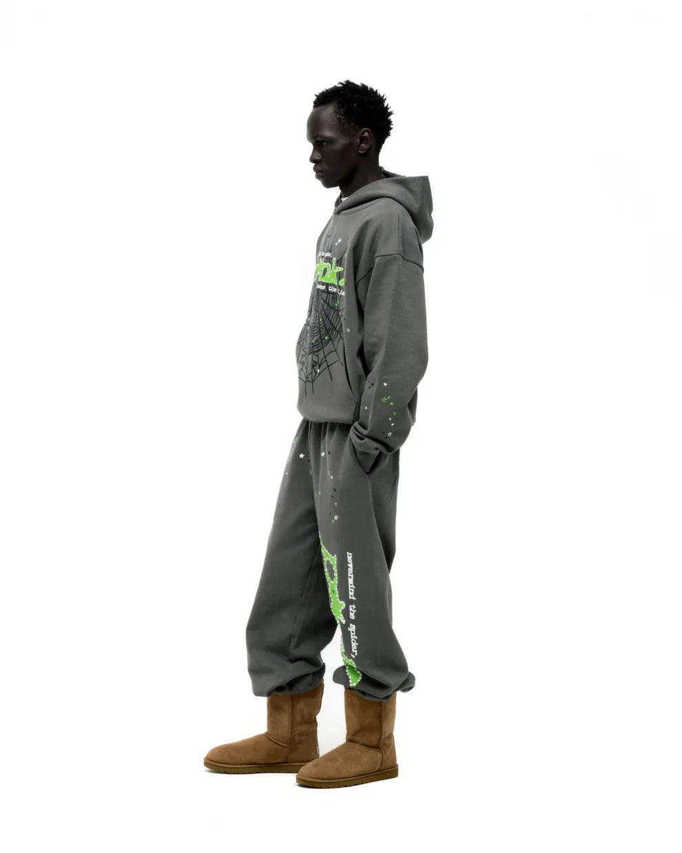 A person stands in profile wearing an olive green Sp5der “P!nk! Hares the Slime” 440GSM heavyweight spider-web pullover hoodie, paired with matching sweatpants and tan boots, showcasing streetwear style against a plain white background.