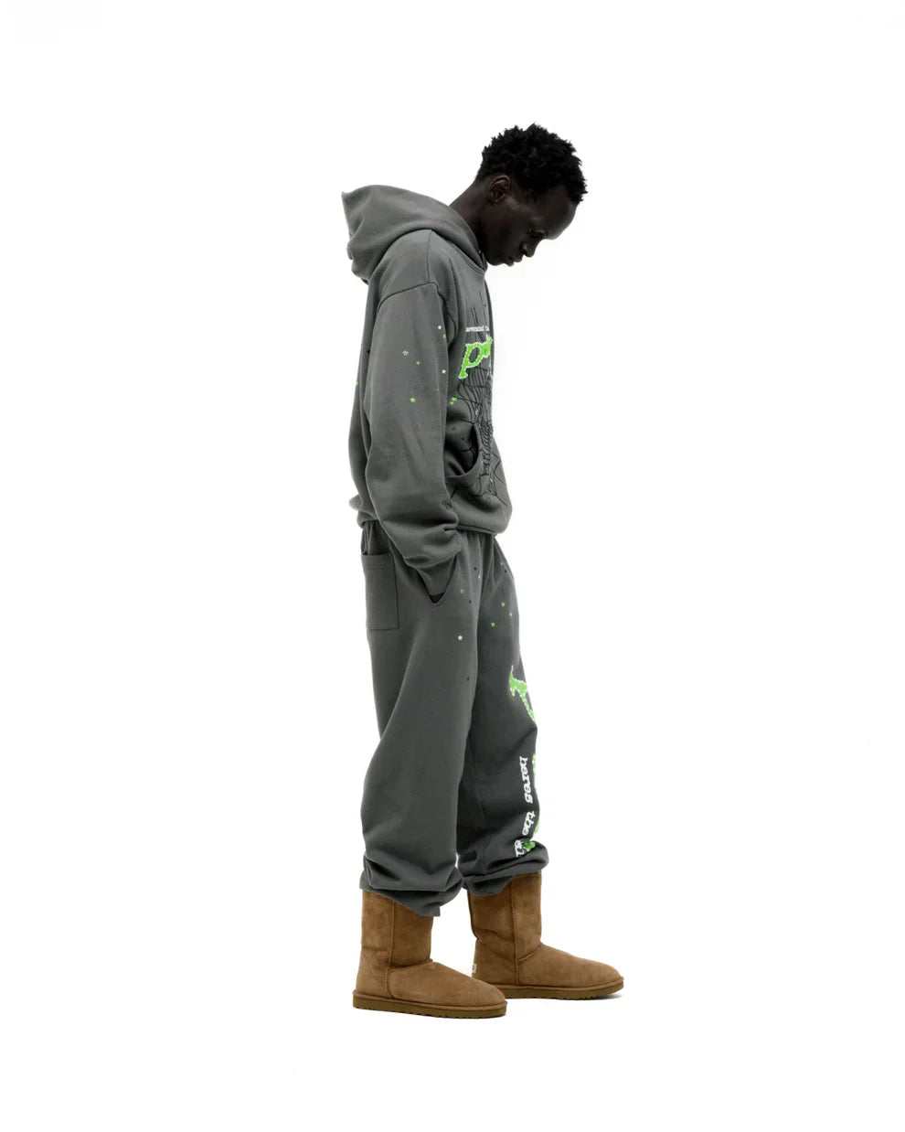A person stands in profile against a white background, wearing an olive green Sp5der “P!nk! Hares the Slime” 440GSM heavyweight spider-web hoodie with their hands in their pockets and head slightly bowed.