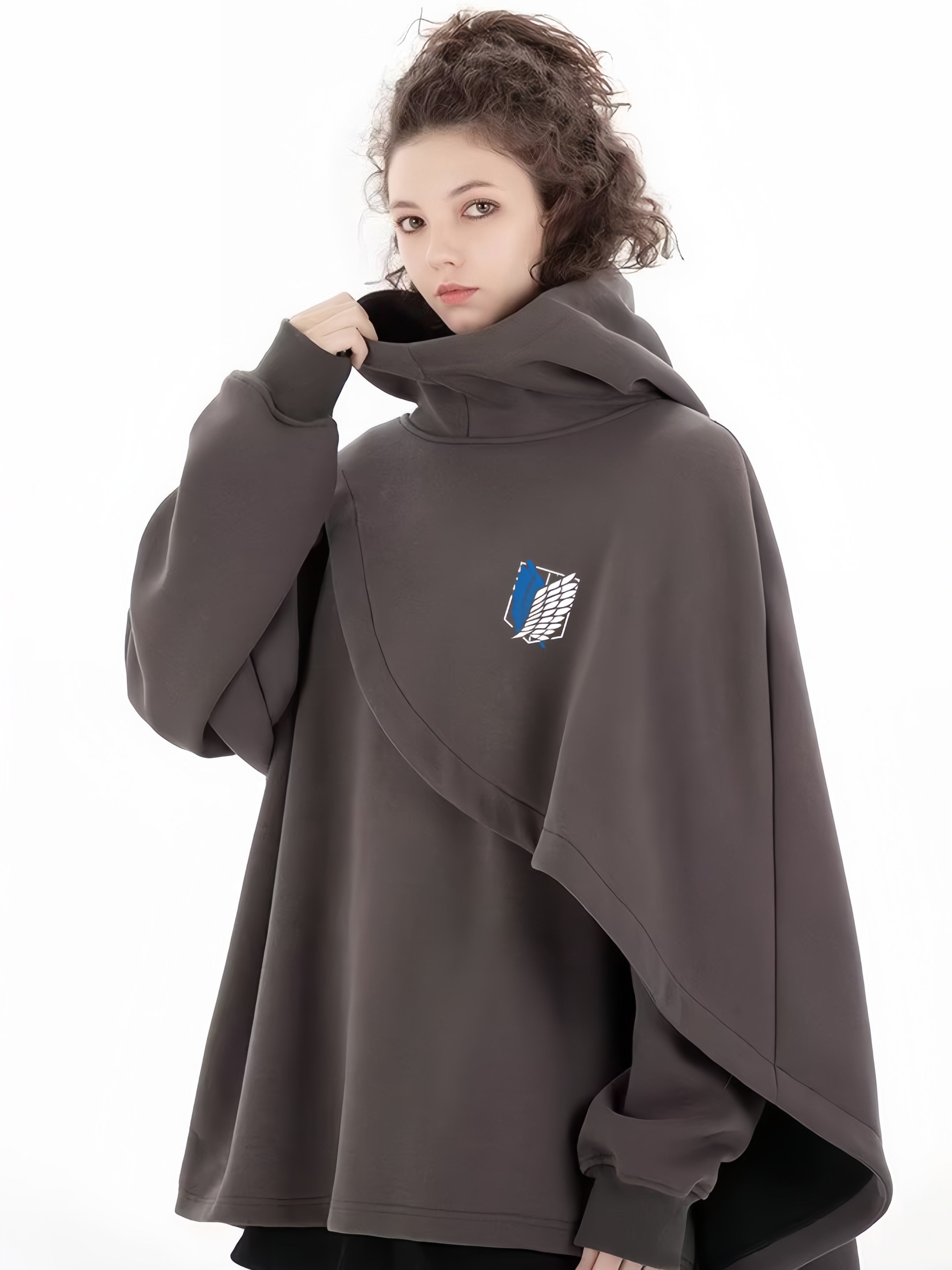 A person with curly hair stands against a white background, wearing an oversized, dark gray Limited Edition Attack on Titan Hooded Cape by Attack on Titan. It features the Survey Corps emblem on the chest and has a high collar they're adjusting.