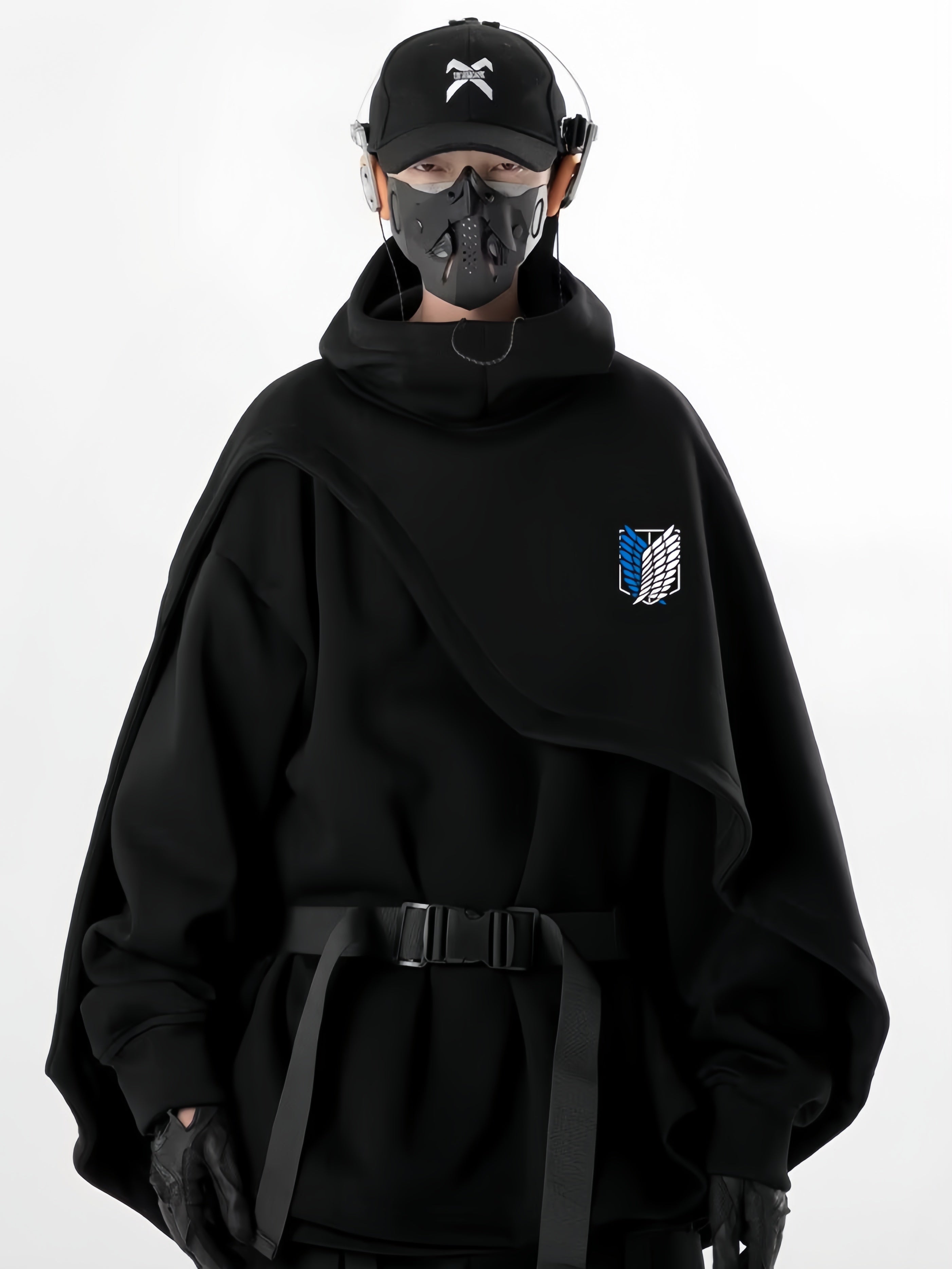 A person in a black futuristic outfit faces forward, featuring the Limited Edition Attack on Titan Hooded Cape - Street Style. Their ensemble includes a mask and cap, with a geometric design patch on their clothing. The background is plain white.