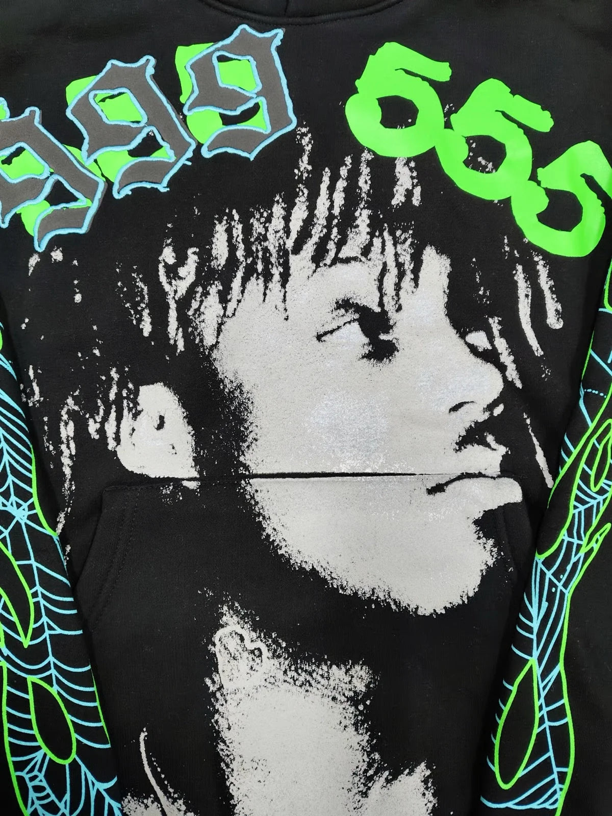 The Sp5der “999 555 Legacy” Hoodie is a black/green/blue 440GSM heavyweight streetwear pullover, featuring a grayscale face graphic, blue "999" and green "666" above, plus blue-green web patterns on the sleeves.