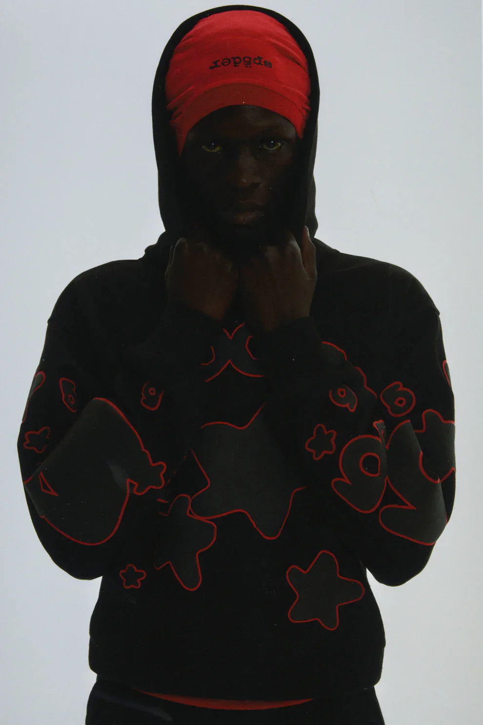 A person in the Sp5der 440GSM Heavyweight Star Hoodie – Black & Grey Oversized Streetwear Pullover stands against a plain background, hood up over a partially hidden red text cap, showcasing bold star designs.