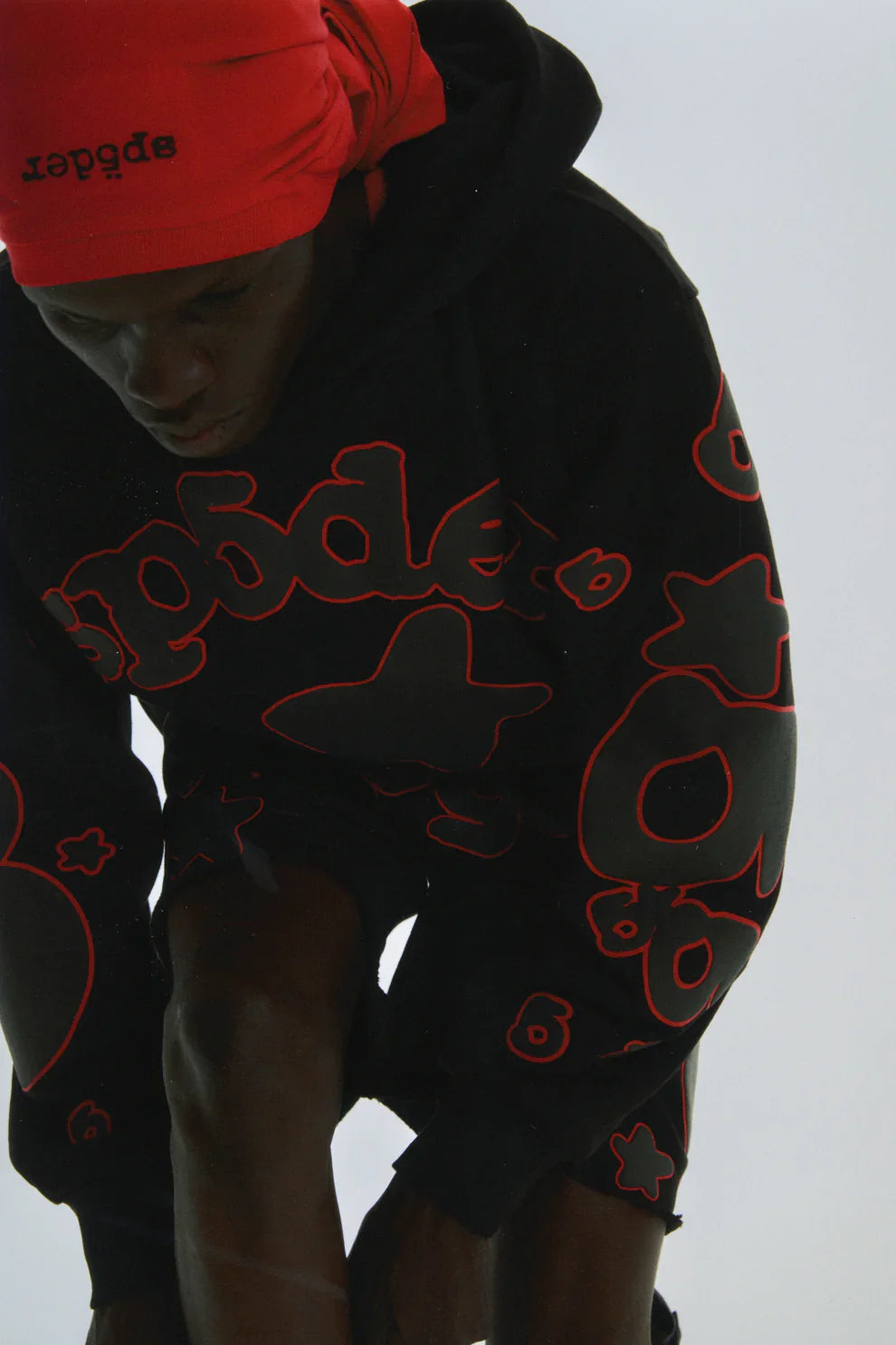 A person in a black Sp5der 440GSM Heavyweight Star Hoodie – Black & Grey Oversized Streetwear Pullover, featuring bold outlined shapes and a red headscarf, looks down against a plain light background—streetwear meets fleece comfort.