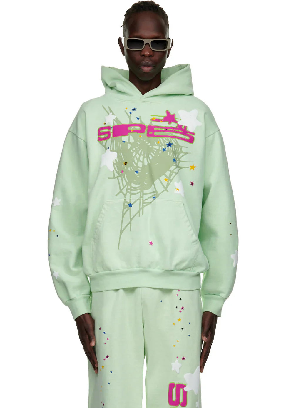 Wearing a Sp5der “Mint Star Web” Hoodie—mint green, 440GSM, oversized—with vibrant graphics like webs and stars plus matching sweatpants channels bold Y2K streetwear energy. The look finishes with dark sunglasses.
