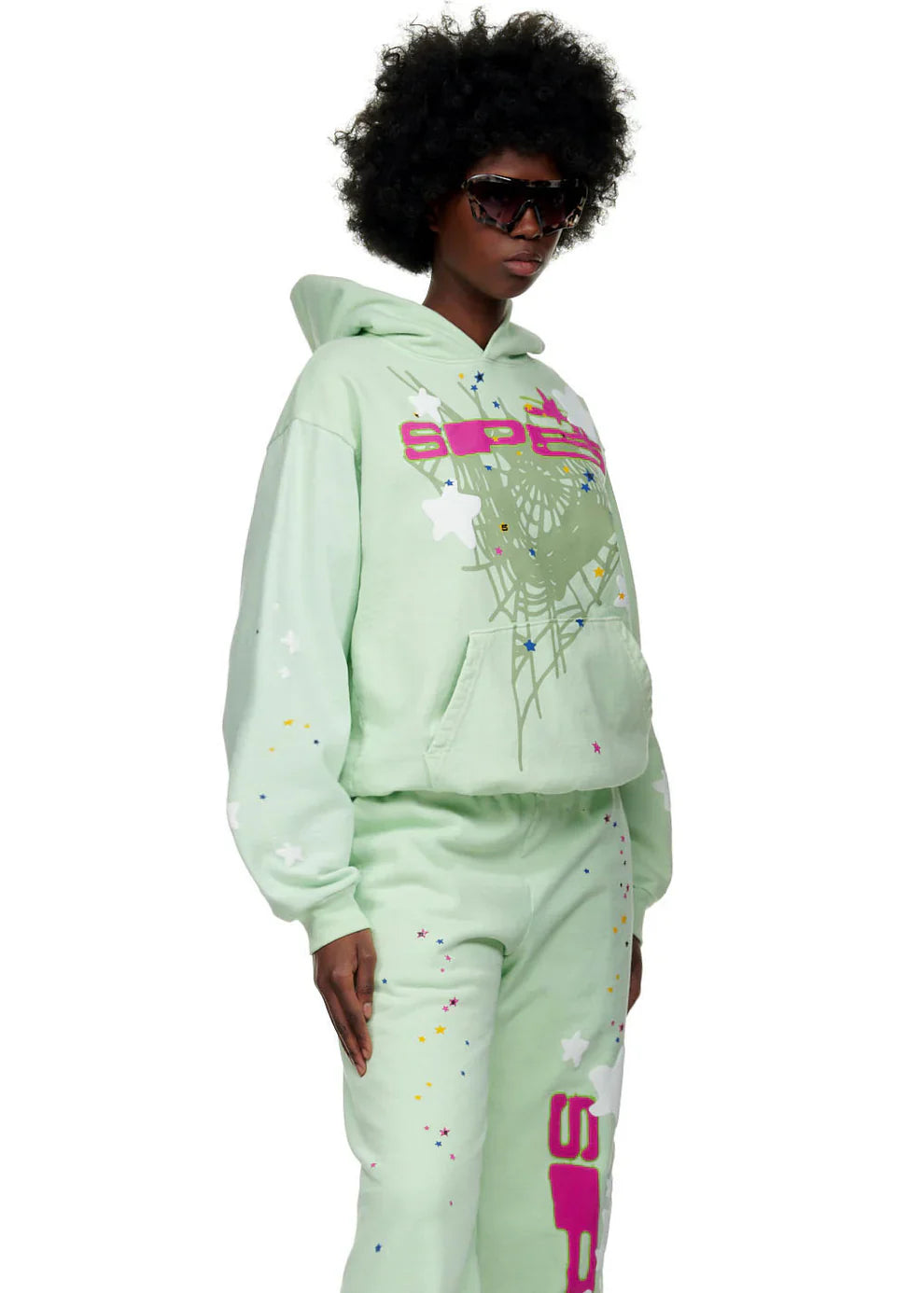 Wearing sunglasses, a person with curly hair rocks the Sp5der “Mint Star Web” Hoodie in mint green—an oversized 440GSM heavyweight streetwear pullover featuring colorful star web graphics, splatters, relaxed fit, and striking Y2K-inspired style.