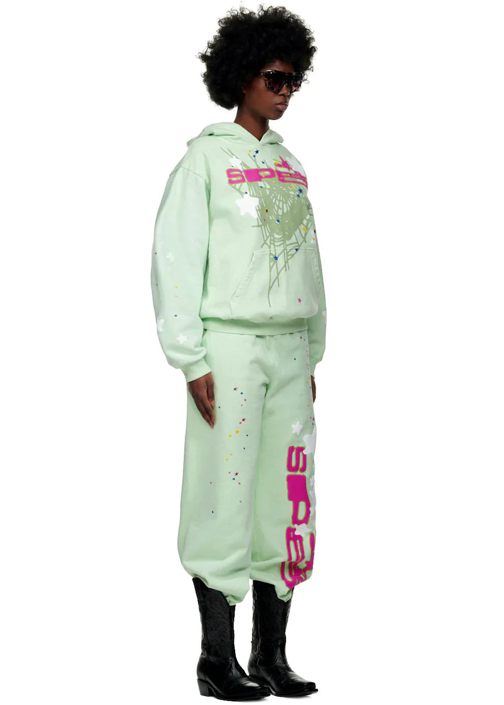 A person in Y2K streetwear—a Sp5der “Mint Star Web” Hoodie (mint green, 440GSM heavyweight oversized pullover), matching splatter sweatpants, sunglasses, and black cowboy boots—stands facing slightly sideways.