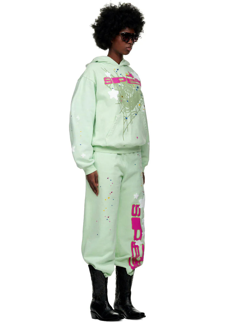A person in Y2K streetwear—a Sp5der “Mint Star Web” Hoodie (mint green, 440GSM heavyweight oversized pullover), matching splatter sweatpants, sunglasses, and black cowboy boots—stands facing slightly sideways.