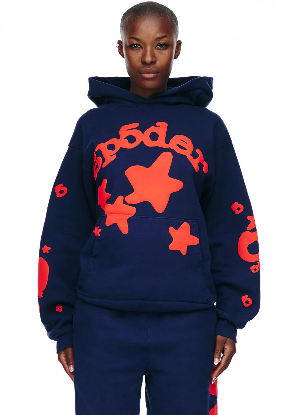 A person wears the Sp5der 440GSM Heavyweight Hoodie in navy blue with bold red star puff print and abstract lettering, paired with matching navy pants. The streetwear pullover pops against a plain white background.
