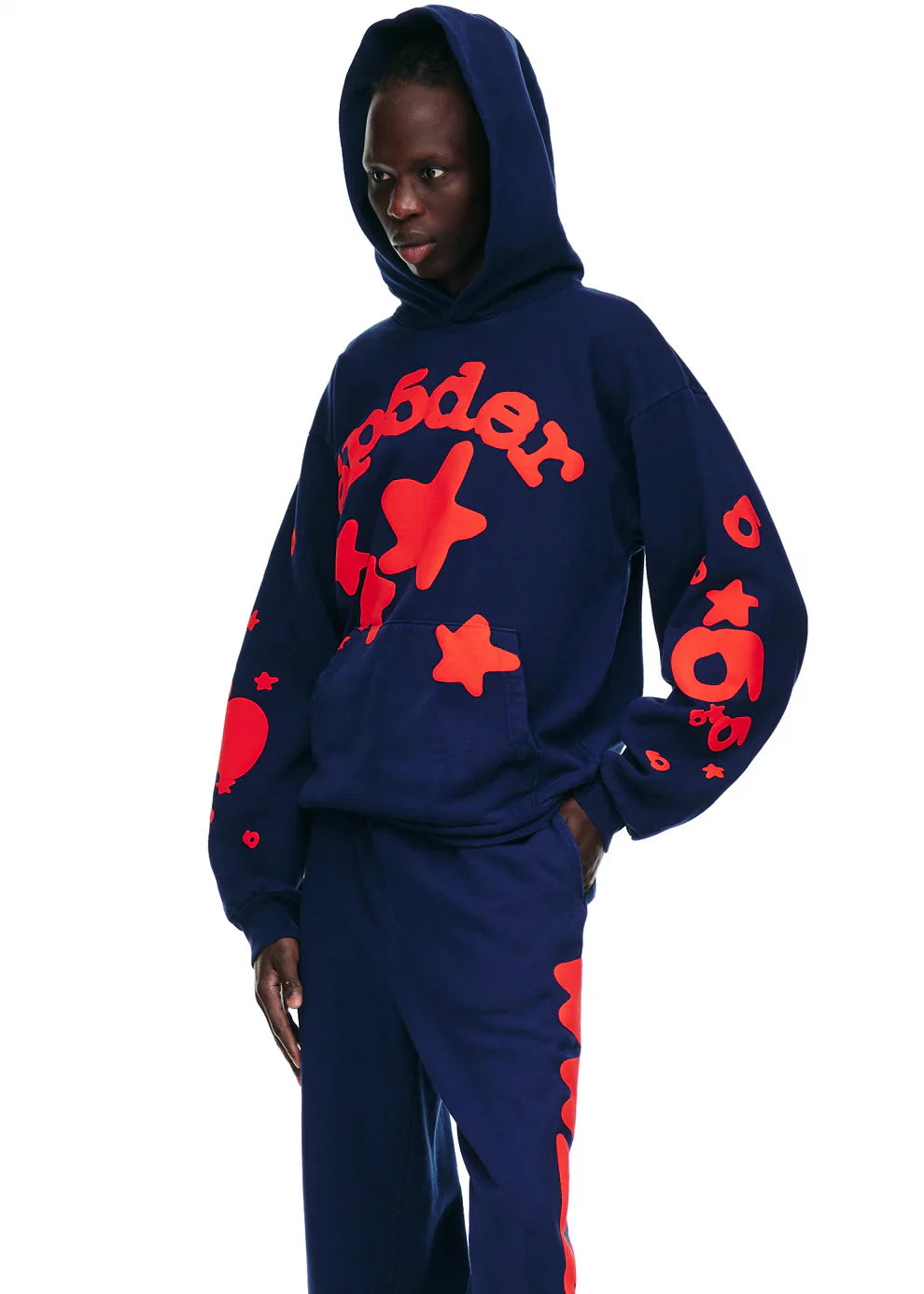 A person models the Sp5der 440GSM Heavyweight Hoodie in navy blue with red star puff print, styled with matching pants, hands in pockets, against a white background for a bold Y2K streetwear vibe.