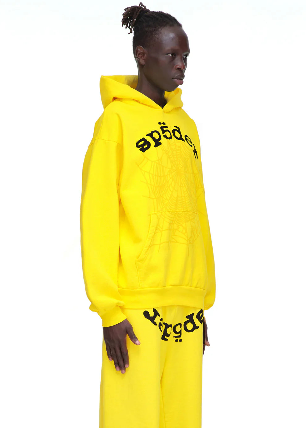 Someone stands against a plain white background wearing the Yellow Graphic Hoodie with Spider Web Print and Lettering – Comfort Fit Pullover Hoodie, paired with matching pants featuring bold black text and spider web designs.