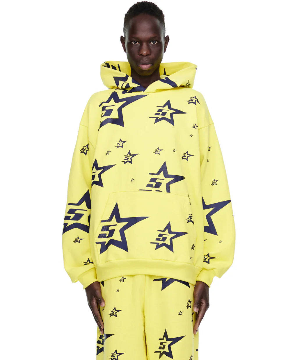 A person wears the Yellow All-Over Star Graphic Hoodie – Comfort Fit Pullover Hoodie, featuring large and small black outlined stars with a stylized E inside, paired with matching pants, standing against a white background.