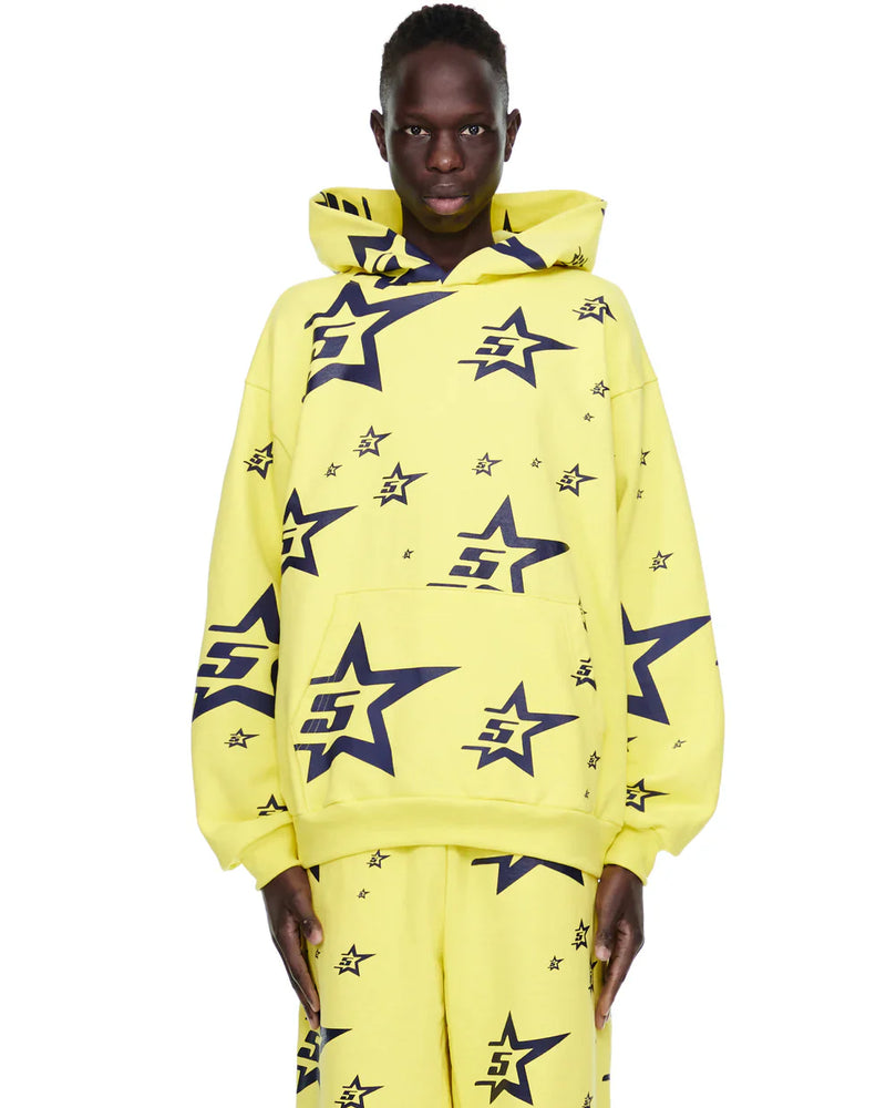 A person wears the Yellow All-Over Star Graphic Hoodie – Comfort Fit Pullover Hoodie, featuring large and small black outlined stars with a stylized E inside, paired with matching pants, standing against a white background.