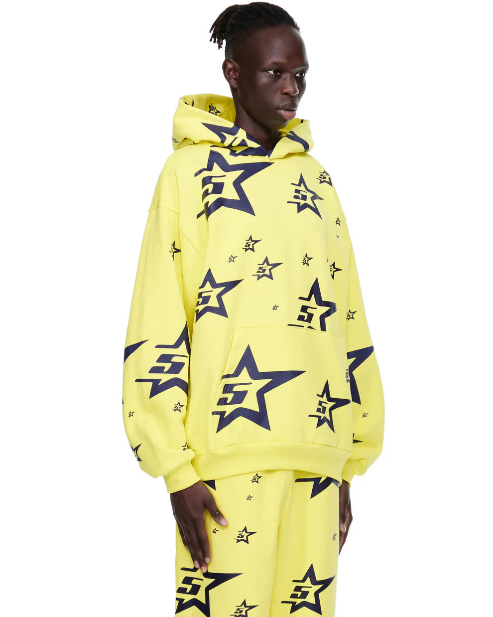 A person stands against a plain white background wearing the Yellow All-Over Star Graphic Hoodie – Comfort Fit Pullover Hoodie and matching pants, both featuring large navy blue outlined stars with S inside some stars.