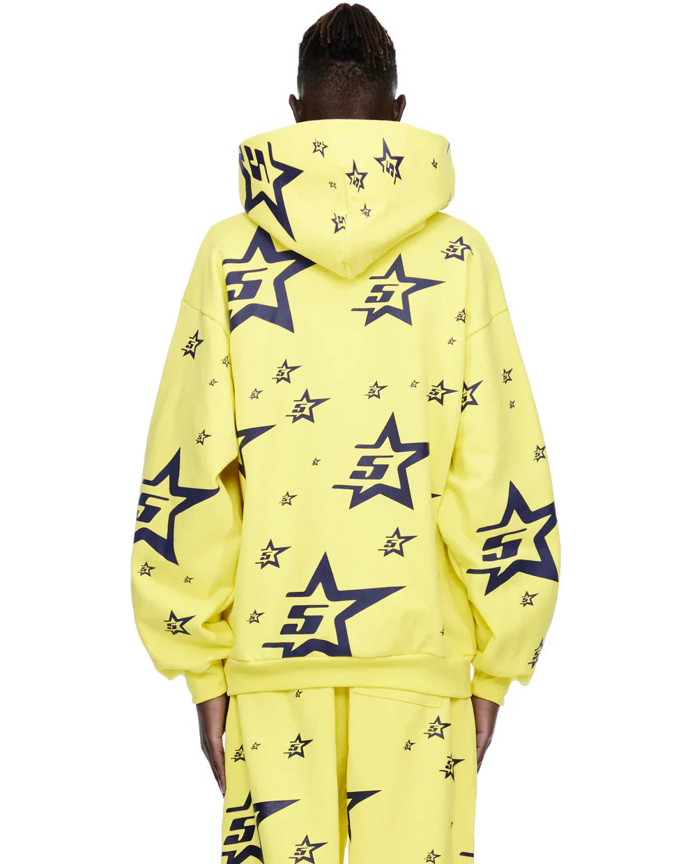 A person faces away from the camera, wearing a Yellow All-Over Star Graphic Hoodie – Comfort Fit Pullover Hoodie with large black outlined stars and small black stars scattered across the fabric, paired with matching pants.
