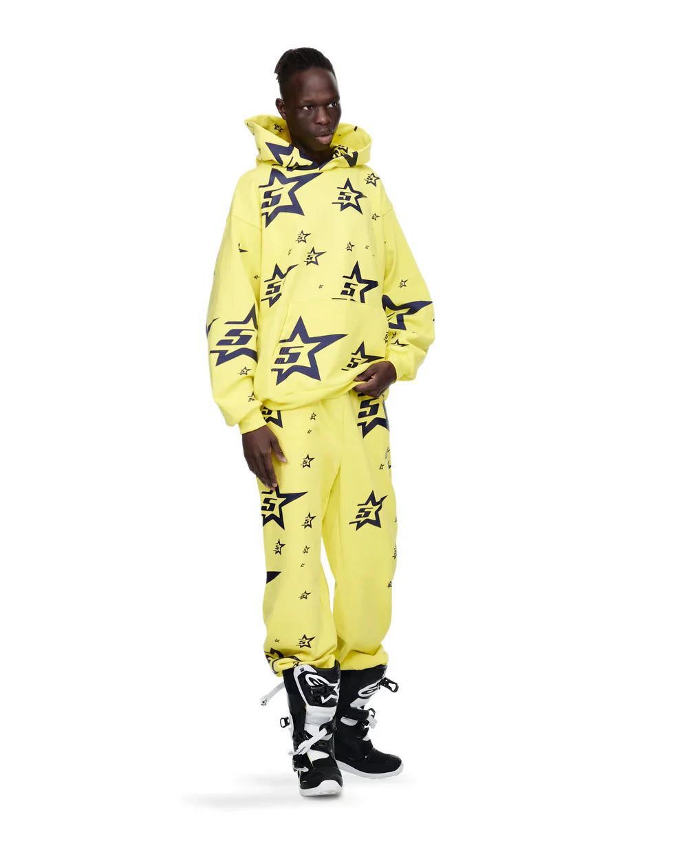 A person wears the Yellow All-Over Star Graphic Hoodie – Comfort Fit Pullover Hoodie with matching star-patterned pants and black-and-white high-top sneakers, standing against a plain white background.