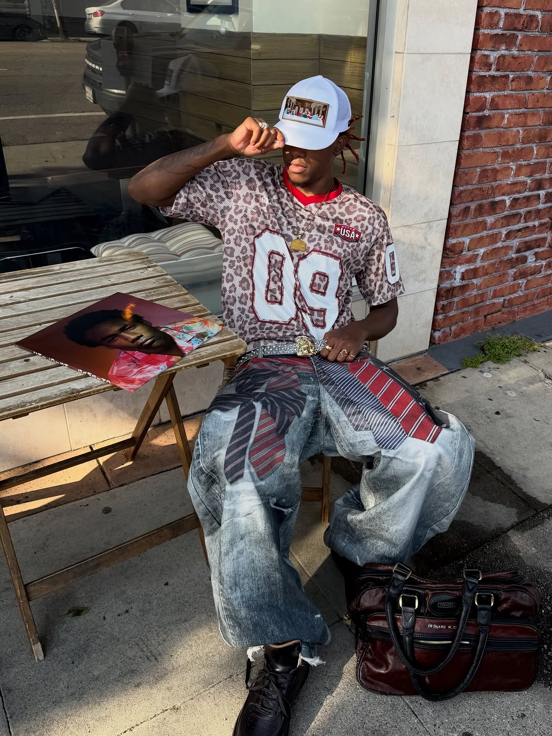 Man seated outdoors wearing a leopard print varsity mesh jersey numbered 09 with wide-leg printed jeans and a white trucker c
