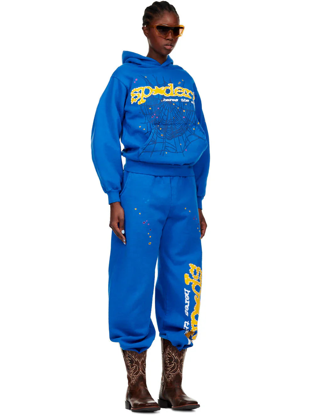 Someone wears a Sp5der “Hares the Slime” Hoodie (440GSM) in royal blue with fuzzy spider-web graphics, matching sweatpants, brown cowboy boots, and large sunglasses, standing against a plain background.