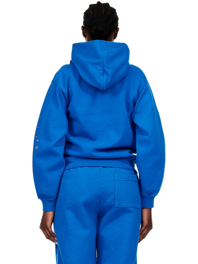A person stands with their back to the camera wearing a Sp5der “Hares the Slime” Hoodie (440GSM) – Royal Blue, featuring a fuzzy logo spider-web graphic and hood up, paired with matching sweatpants.