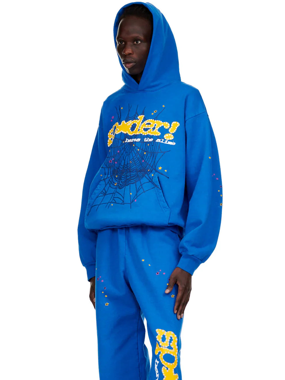 A person in a royal blue Sp5der “Hares the Slime” Hoodie (440GSM) with a fuzzy yellow and white spider-web logo, styled with matching sweatpants, stands against a plain white background.