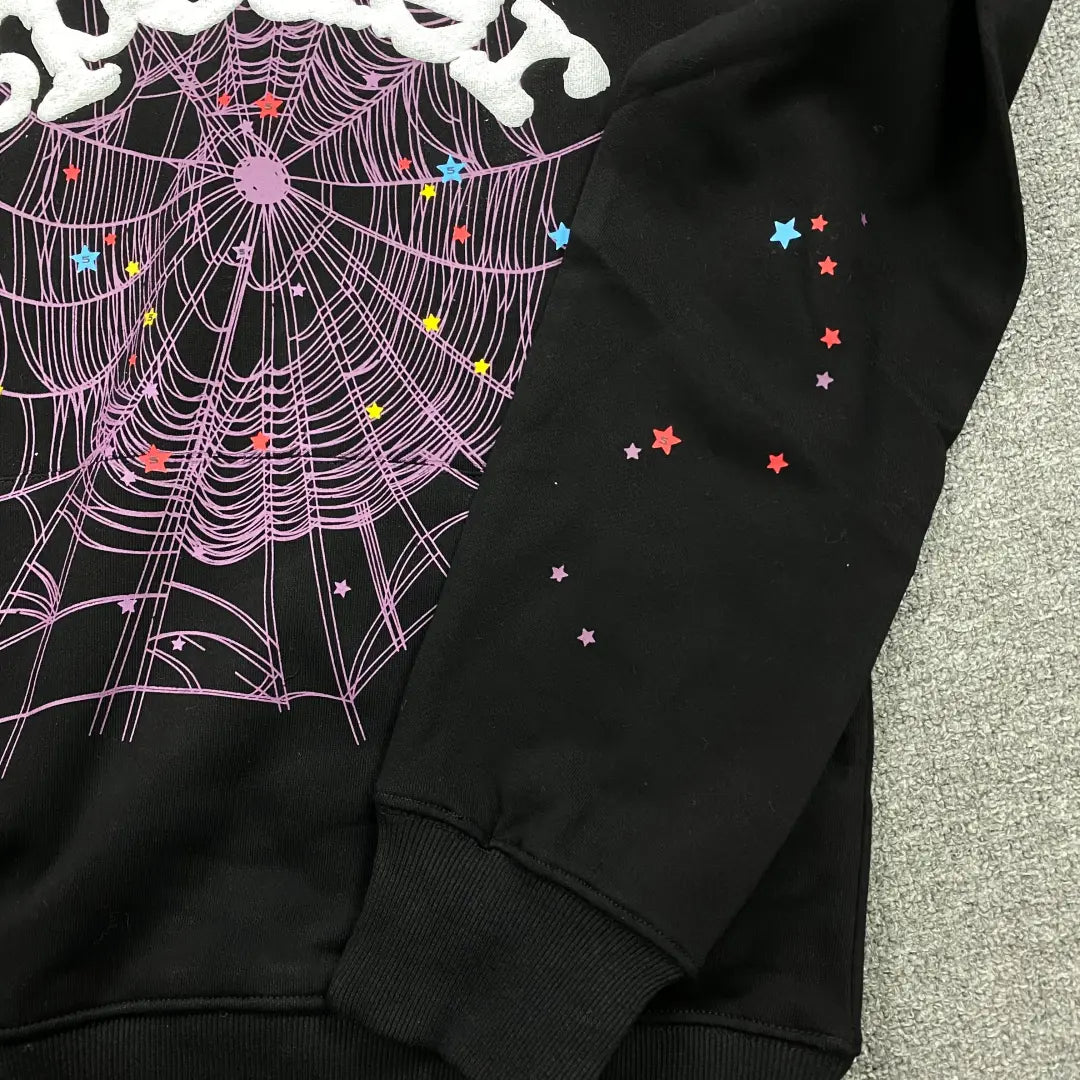 The Black Sp5der Web Hoodie 440GSM features a large purple web and white star accents on the front and sleeve, displayed on a gray textured surface.