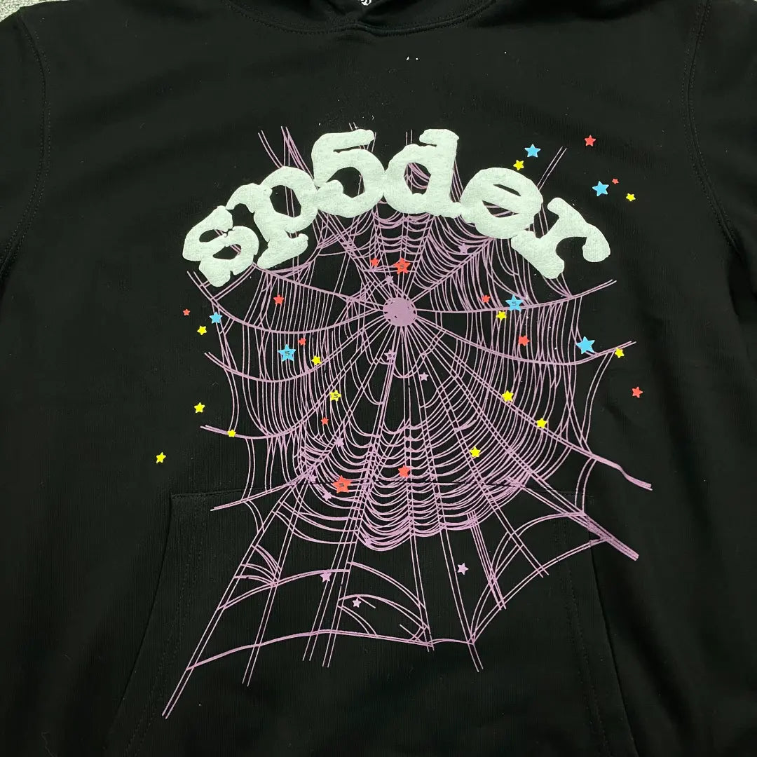 The Black Sp5der Web Hoodie 440GSM features bold white “sp5der” text above a large purple web and scattered white stars, all on a black hoodie.