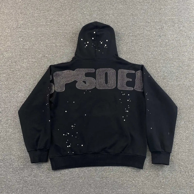A black Sp5der Black Rhinestone Spiderweb Crystal Hoodie lies flat on gray, its back adorned with SPACE in large dotted letters and scattered white dots like stars, capturing the bold streetwear style.