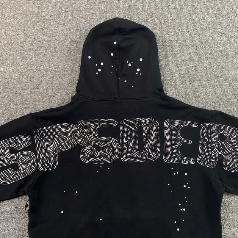 The Sp5der Black Rhinestone Spiderweb Crystal Hoodie features SP5DER in large silver-studded letters on the back and white spots on black fabric, laid flat on a gray surface—a bold streetwear essential.
