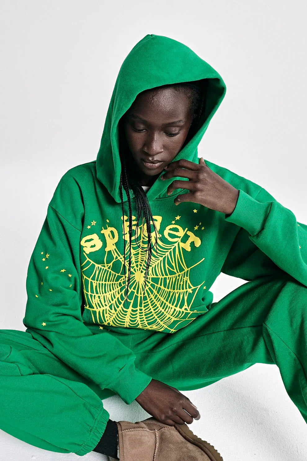 A person in the Sp5der Web Hoodie – Emerald Green & Yellow 440GSM Heavyweight Oversized Streetwear Pullover sits on the floor, hood up, hands on legs. The bold yellow “spider” text and web graphic capture a Y2K streetwear vibe.