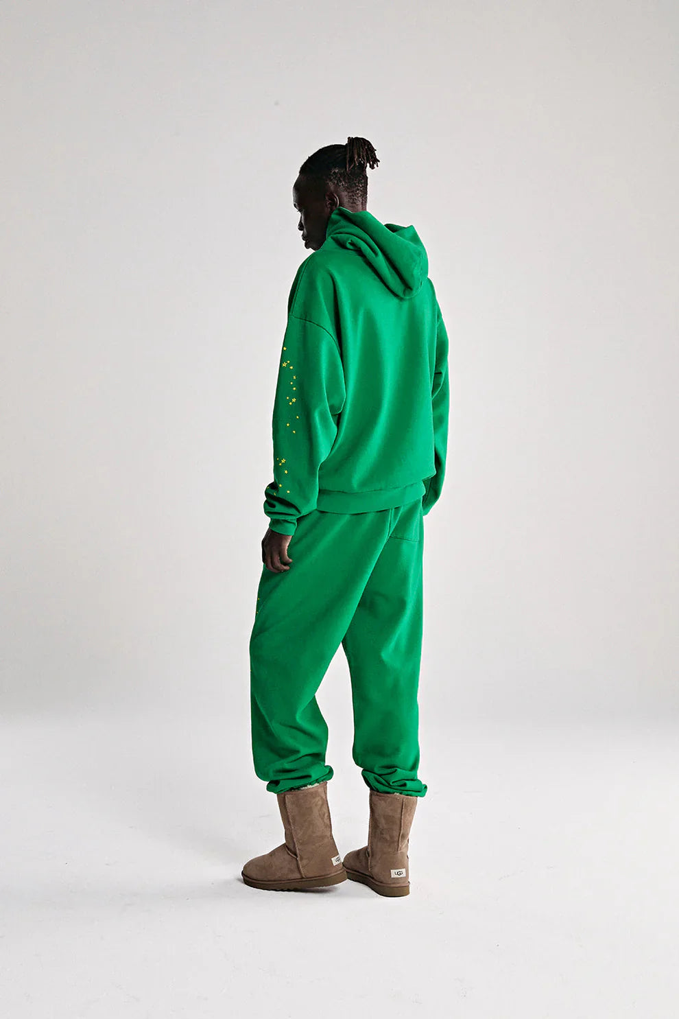 A person faces away in the Sp5der Web Hoodie – Emerald Green & Yellow 440GSM Heavyweight Oversized Streetwear Pullover with matching pants, yellow sleeve dots, and light brown boots, capturing Y2K streetwear vibes on a plain white background.