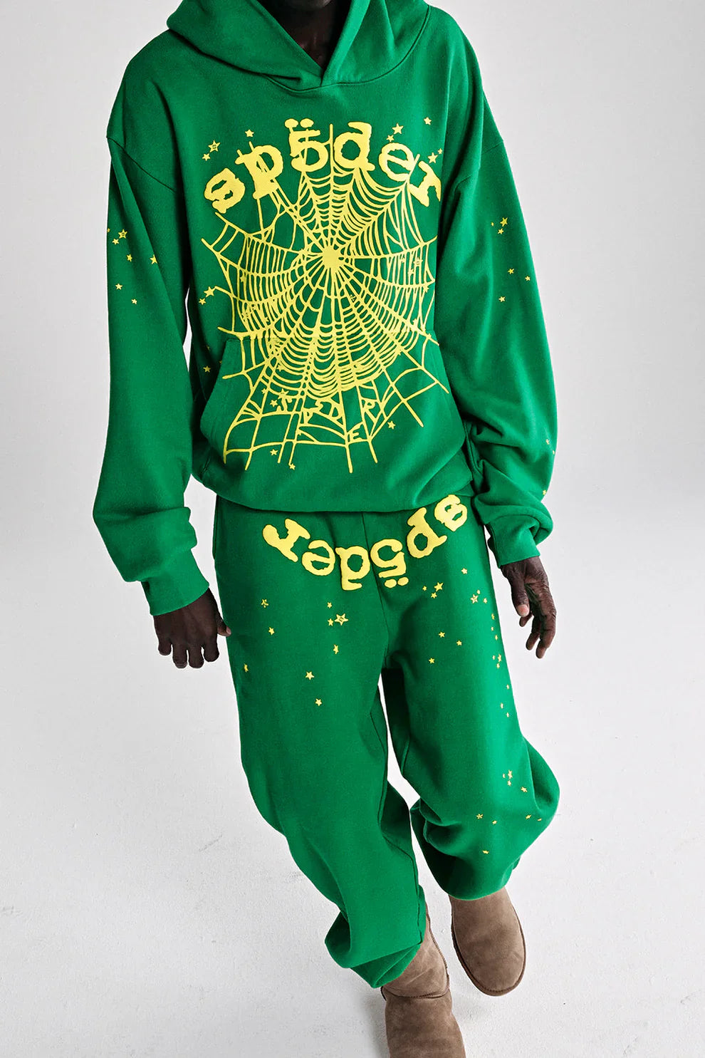 A person in the Sp5der Web Hoodie – Emerald Green & Yellow 440GSM Heavyweight Oversized Streetwear Pullover with matching sweatpants, yellow spiderwebs, and stars paired with brown boots poses against a plain background, channeling Y2K streetwear vibes.