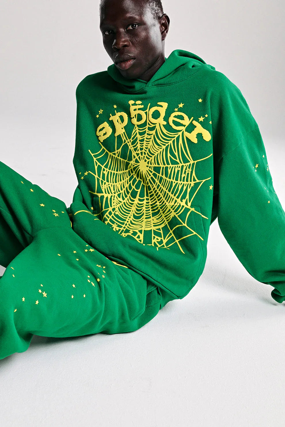Wearing the Sp5der Web Hoodie – Emerald Green & Yellow, a person sits on a white floor against a plain background, showcasing bold Y2K streetwear style with oversized fit and signature yellow star and web designs.