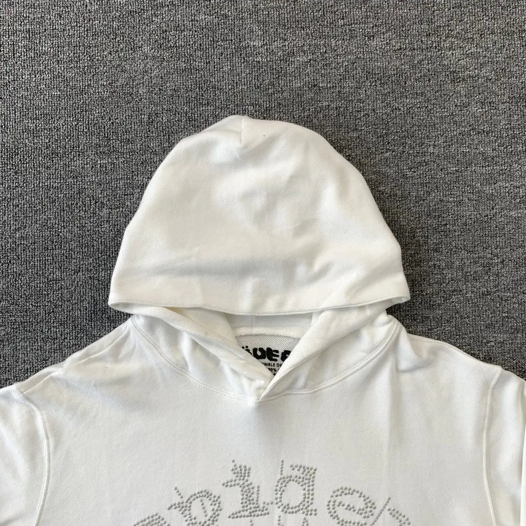 The White Sp5der Crystal Web Hoodie 440GSM features a white rhinestone spider web design on the chest. Laid flat on a gray surface with the hood up, it showcases luxury streetwear style and part of the neck tag peeks out inside.