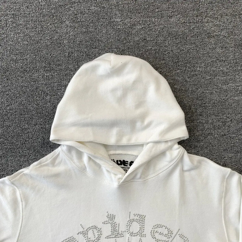 The White Sp5der Crystal Web Hoodie 440GSM features a white rhinestone spider web design on the chest. Laid flat on a gray surface with the hood up, it showcases luxury streetwear style and part of the neck tag peeks out inside.