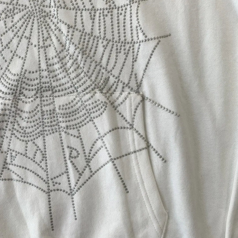 Close-up of the White Sp5der Crystal Web Hoodie 440GSM, showing its white fabric with a crystal rhinestone spiderweb pattern inspired by luxury streetwear trends.