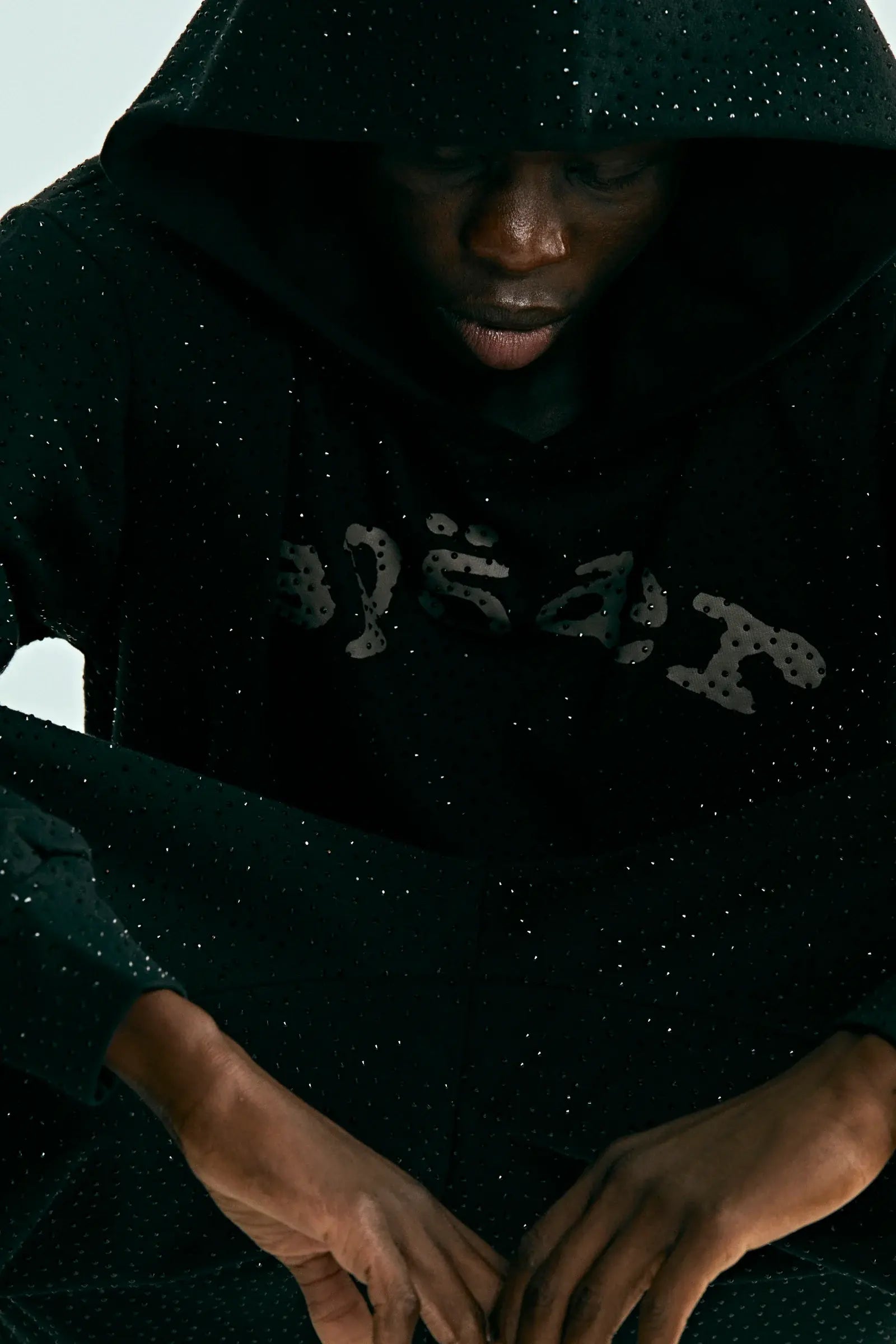 A person wearing the Black Sp5der Rhinestone Logo VVS Hoodie 440GSM – Black Crystal Sp5der VVS Hoodie All-Over Rhinestones, hood up and head down, partially obscures their face as their hands rest on their knees.