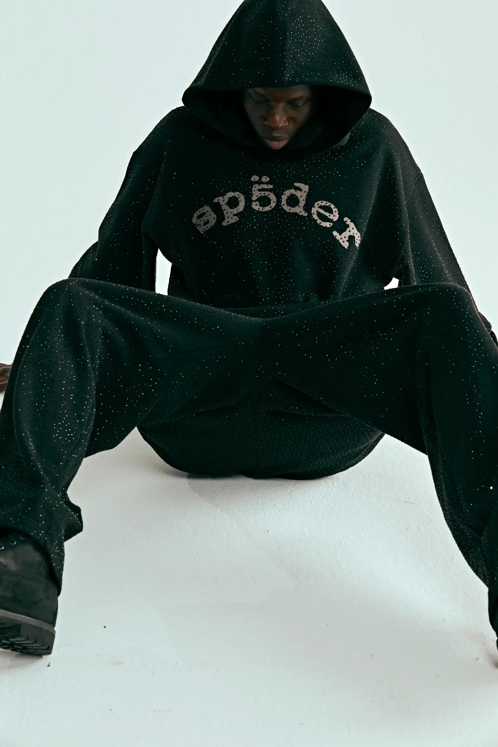 A person in the Black Sp5der Rhinestone Logo VVS Hoodie 440GSM – Black Crystal Sp5der VVS Hoodie All-Over Rhinestones and matching pants sits on the floor, hood up and face partly shadowed, sparkling with rhinestone details.