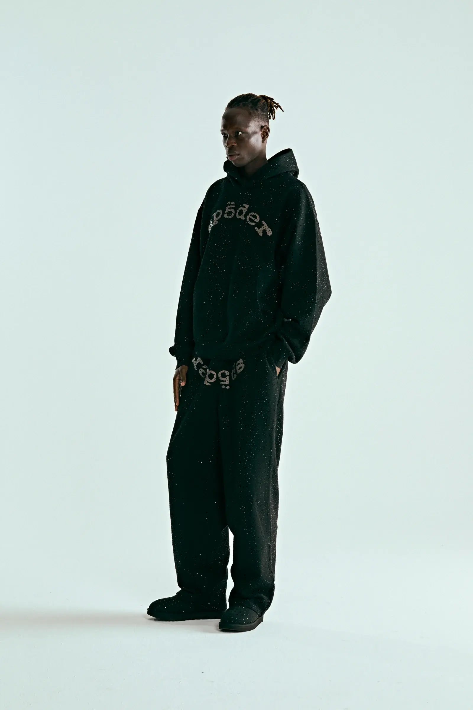 A person stands against a plain white background wearing the Black Sp5der Rhinestone Logo VVS Hoodie 440GSM—Black Crystal Sp5der VVS Hoodie with all-over rhinestones—styled with matching loose, comfortable black pants featuring text designs.
