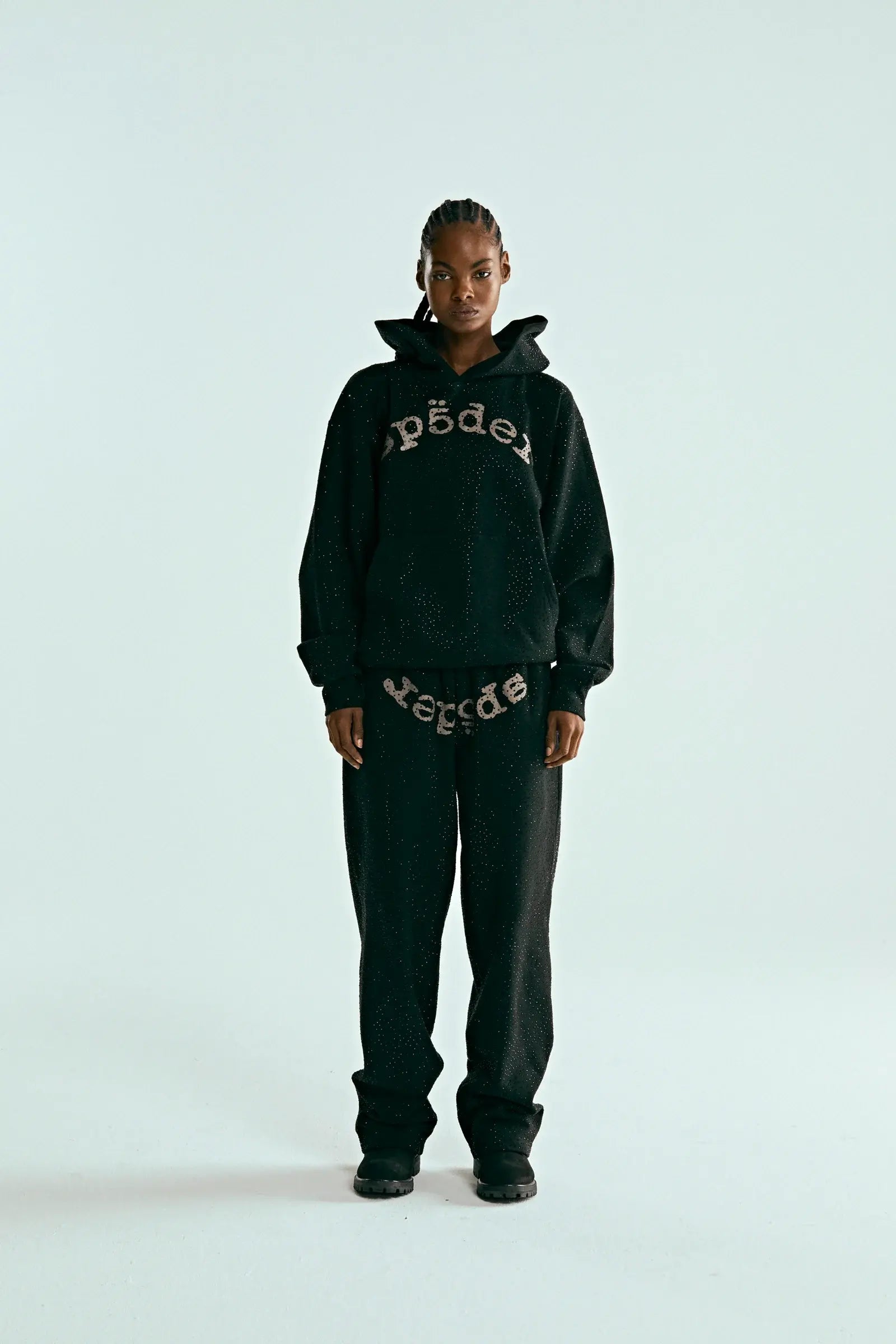 A person with braided hair stands against a plain light background, wearing the Black Sp5der Rhinestone Logo VVS Hoodie 440GSM – Black Crystal Sp5der VVS Hoodie All-Over Rhinestones, matching black pants, and black shoes.