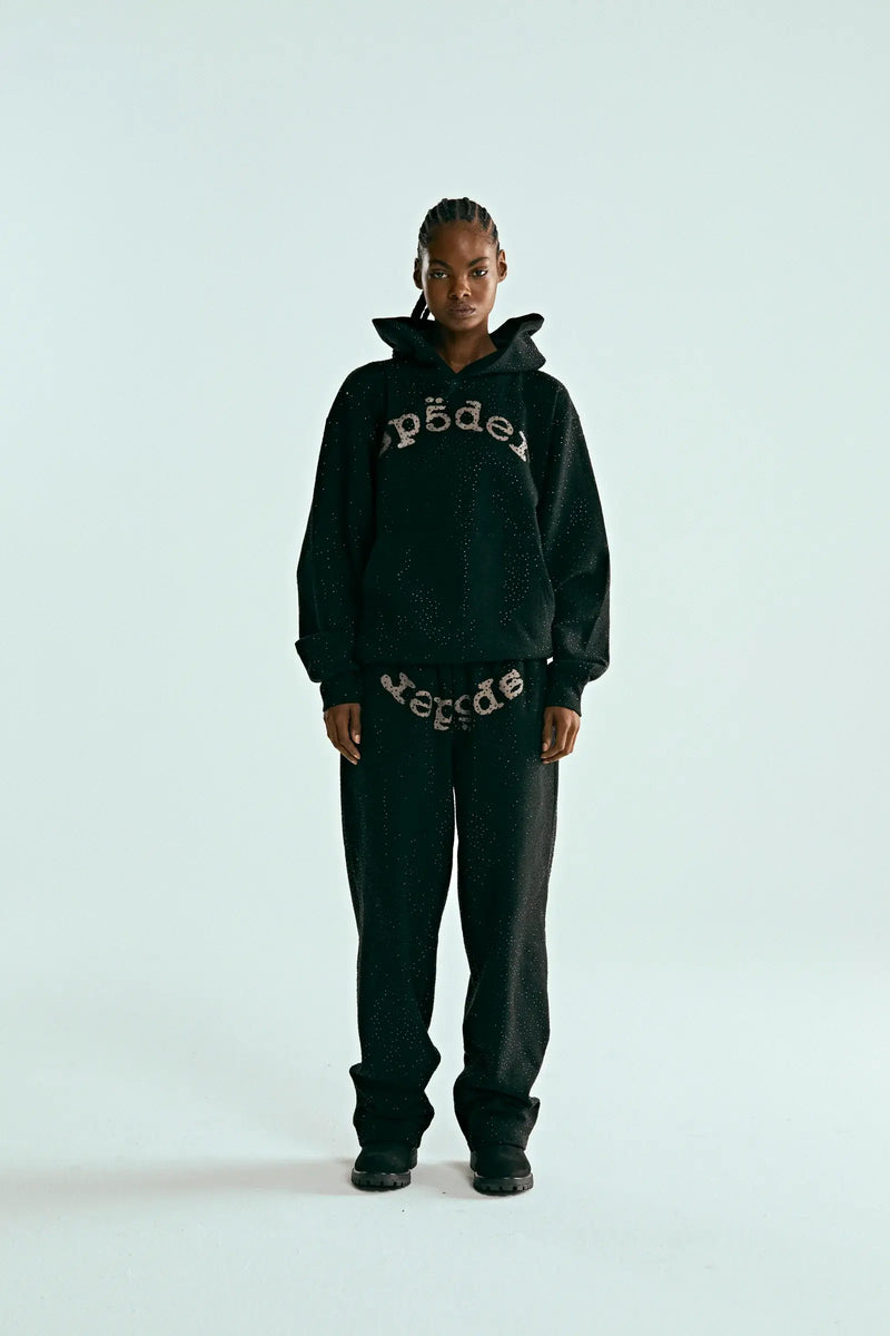 A person with braided hair stands against a plain light background, wearing the Black Sp5der Rhinestone Logo VVS Hoodie 440GSM – Black Crystal Sp5der VVS Hoodie All-Over Rhinestones, matching black pants, and black shoes.