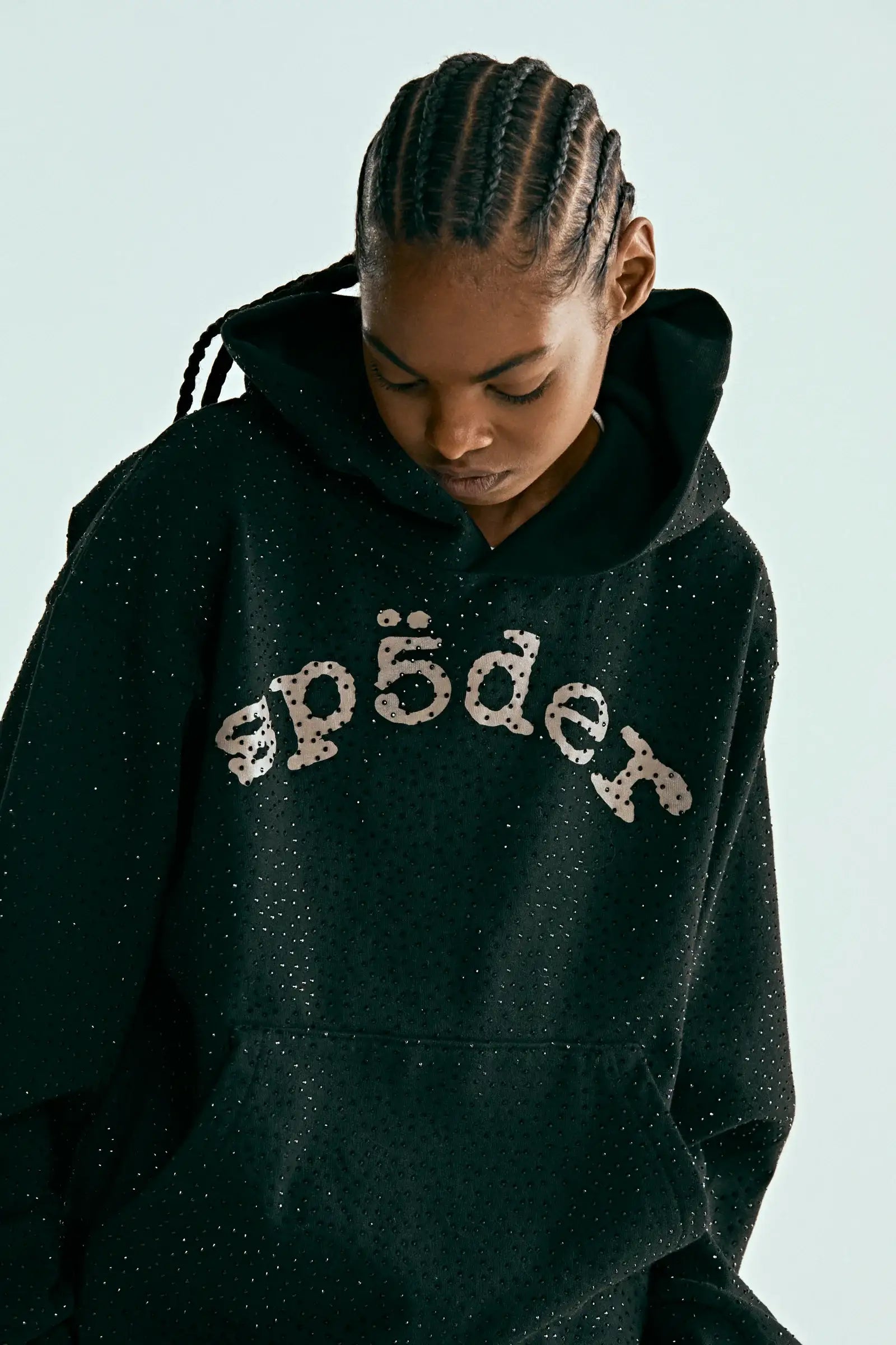 A person with braided hair looks down while wearing the Black Sp5der Rhinestone Logo VVS Hoodie 440GSM, which features all-over rhinestones and "sp5der" in white stylized text across the front.