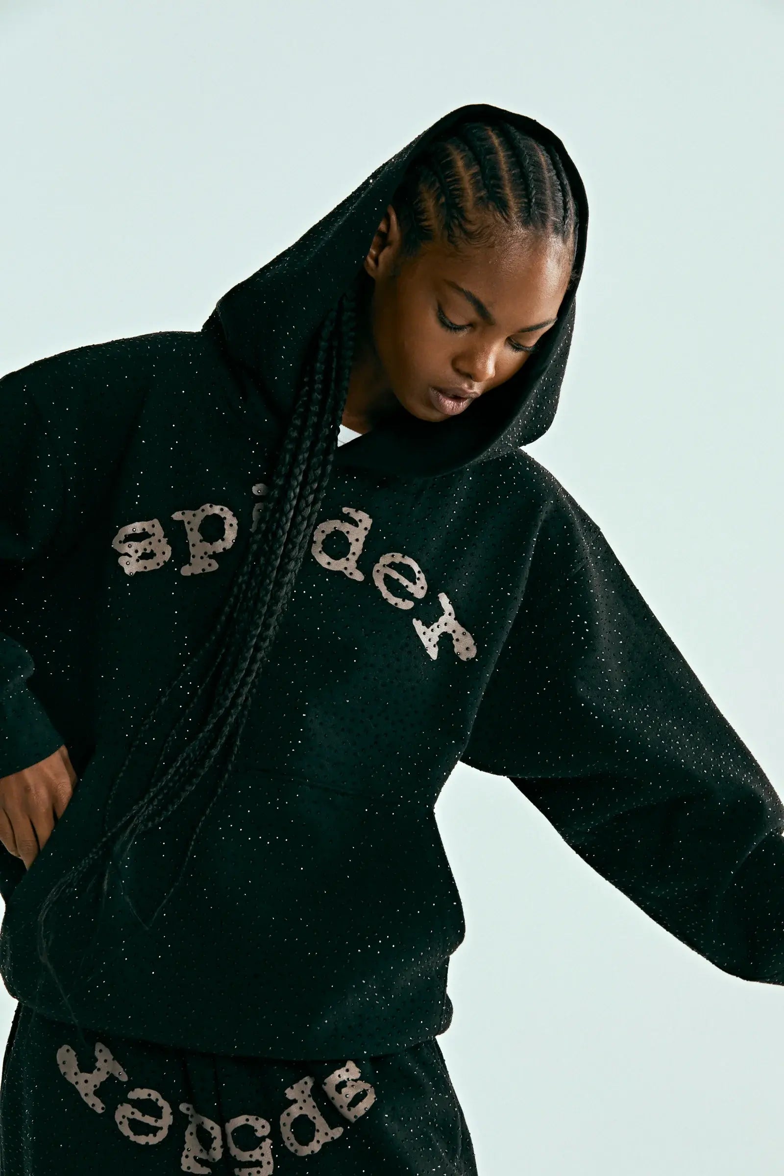 Wearing the Black Sp5der Rhinestone Logo VVS Hoodie 440GSM with matching pants, both featuring sp5der prints and all-over rhinestones, the person looks down as their long braided hair falls forward and one arm is raised.