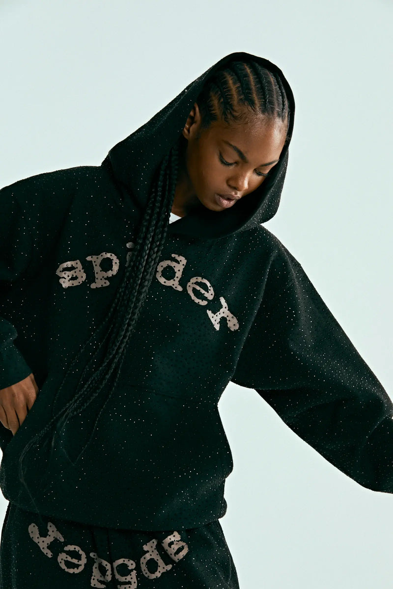 Wearing the Black Sp5der Rhinestone Logo VVS Hoodie 440GSM with matching pants, both featuring sp5der prints and all-over rhinestones, the person looks down as their long braided hair falls forward and one arm is raised.