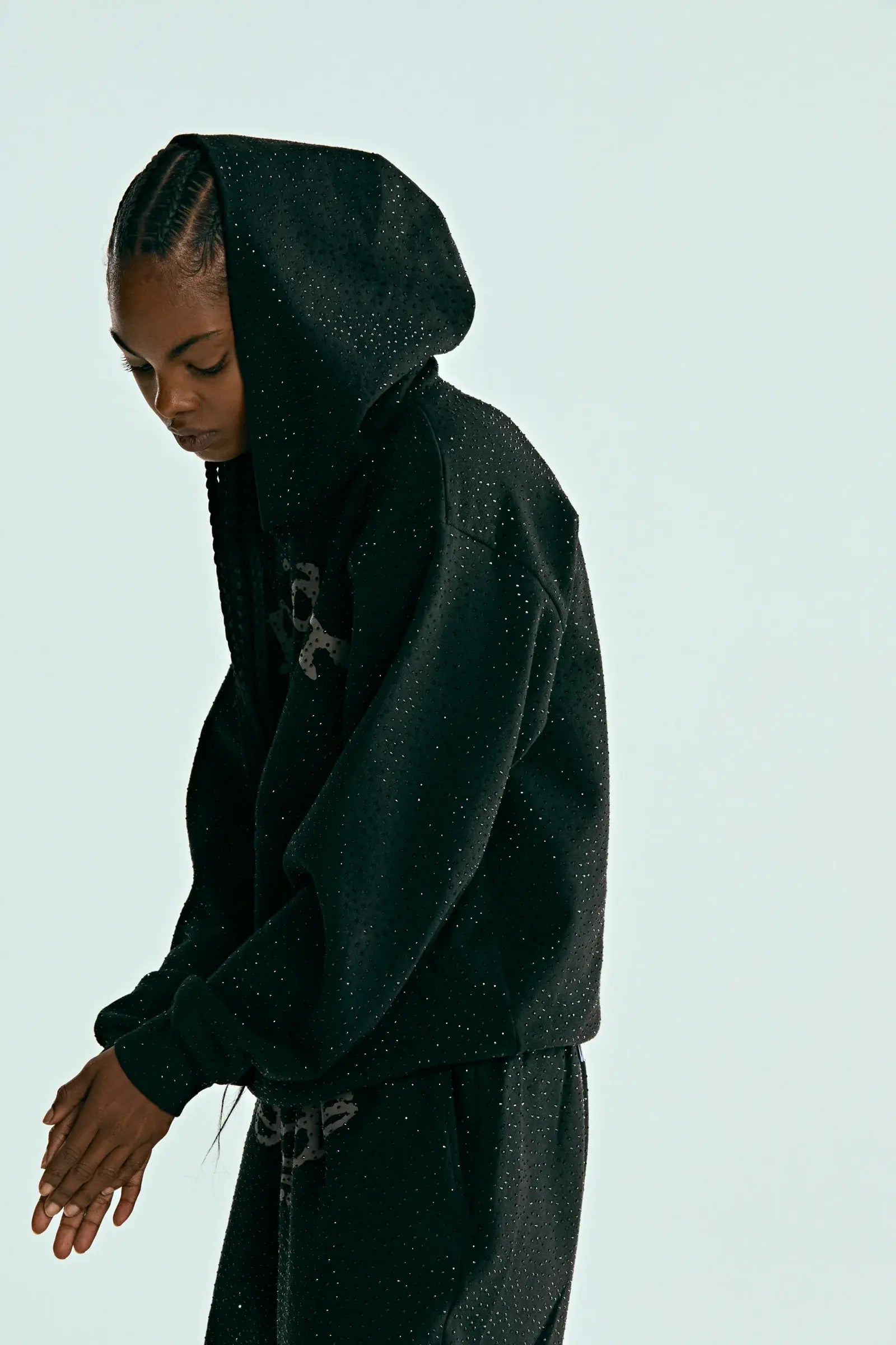 A person wears the Black Sp5der Rhinestone Logo VVS Hoodie 440GSM – Black Crystal Sp5der VVS Hoodie All-Over Rhinestones and matching pants against a light backdrop, looking down with hands together. The outfit features subtle rhinestone accents.