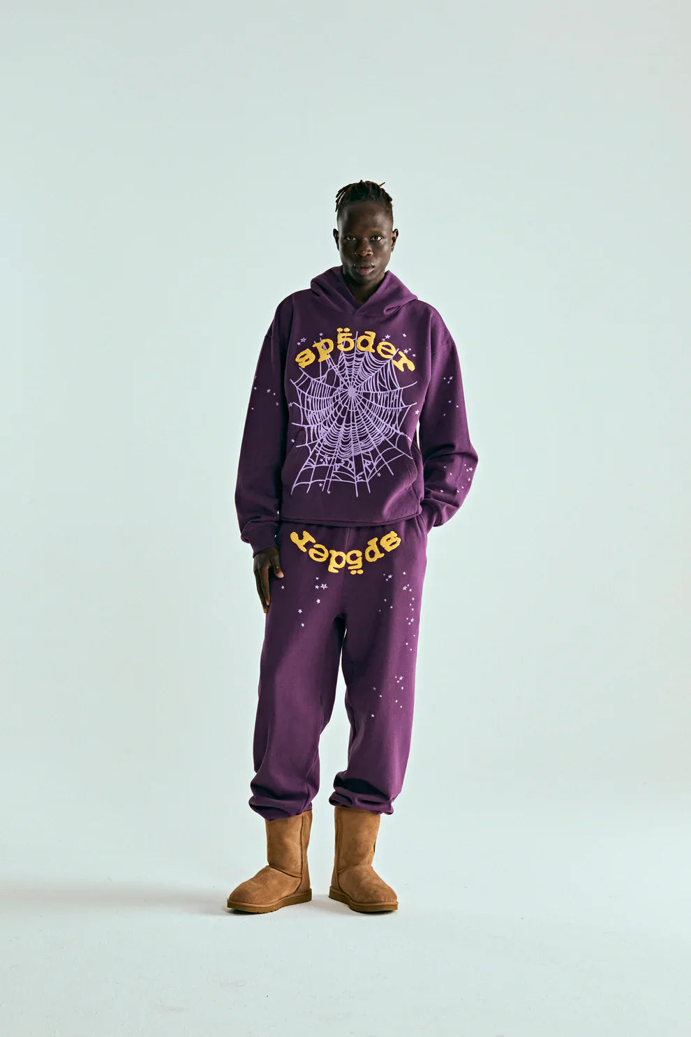 A person stands against a plain background wearing the Sp5der Web Hoodie – Deep Purple & Yellow 440GSM Heavyweight Oversized Streetwear Pullover, sweatpants, and tan boots—ideal for luxury streetwear and Y2K fashion fans.