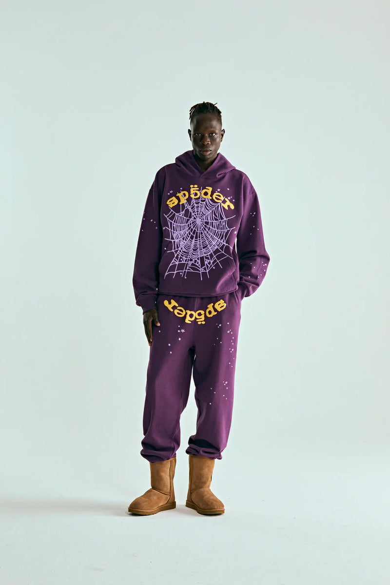 A person stands against a plain background wearing the Sp5der Web Hoodie – Deep Purple & Yellow 440GSM Heavyweight Oversized Streetwear Pullover, sweatpants, and tan boots—ideal for luxury streetwear and Y2K fashion fans.