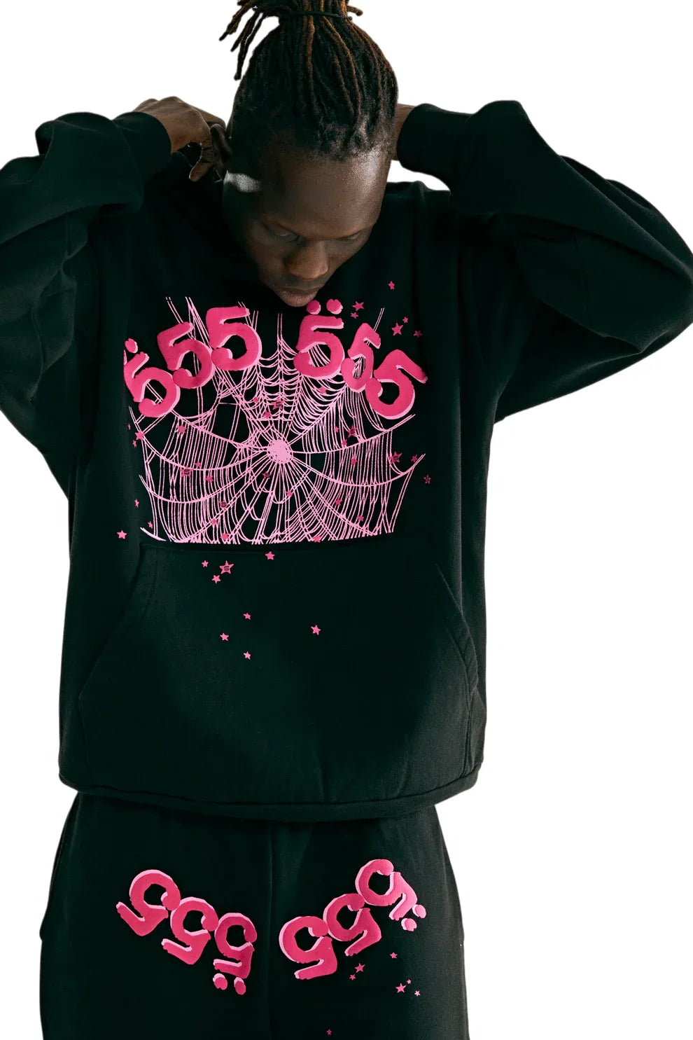A person looks down while wearing a Sp5der 555 Hoodie – Black & Pink Web 440GSM Heavyweight Streetwear Pullover and matching pants, featuring bright pink "555" and "999" designs, spiderwebs, and stars, reflecting Y2K style on a white background.