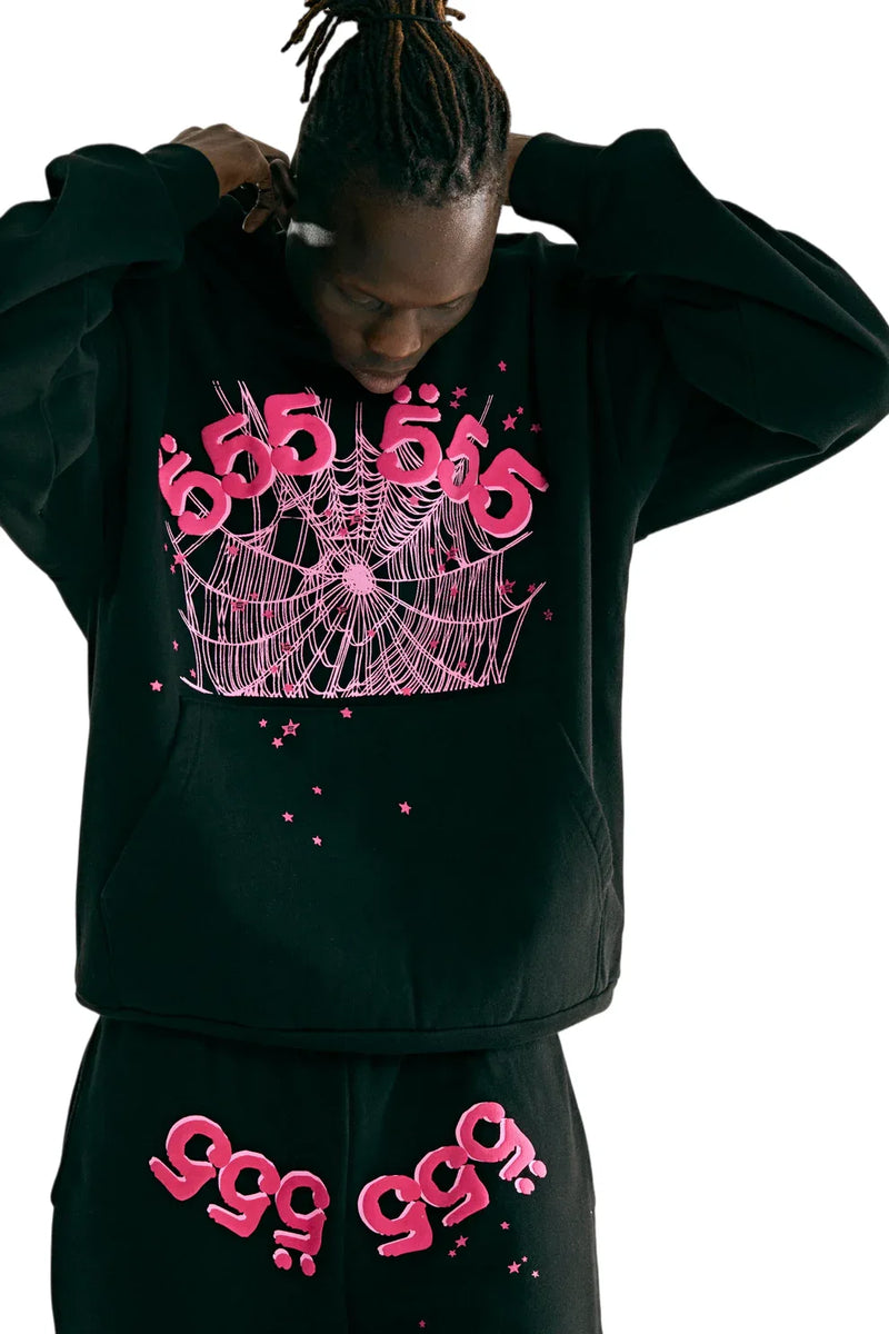 A person looks down while wearing a Sp5der 555 Hoodie – Black & Pink Web 440GSM Heavyweight Streetwear Pullover and matching pants, featuring bright pink "555" and "999" designs, spiderwebs, and stars, reflecting Y2K style on a white background.